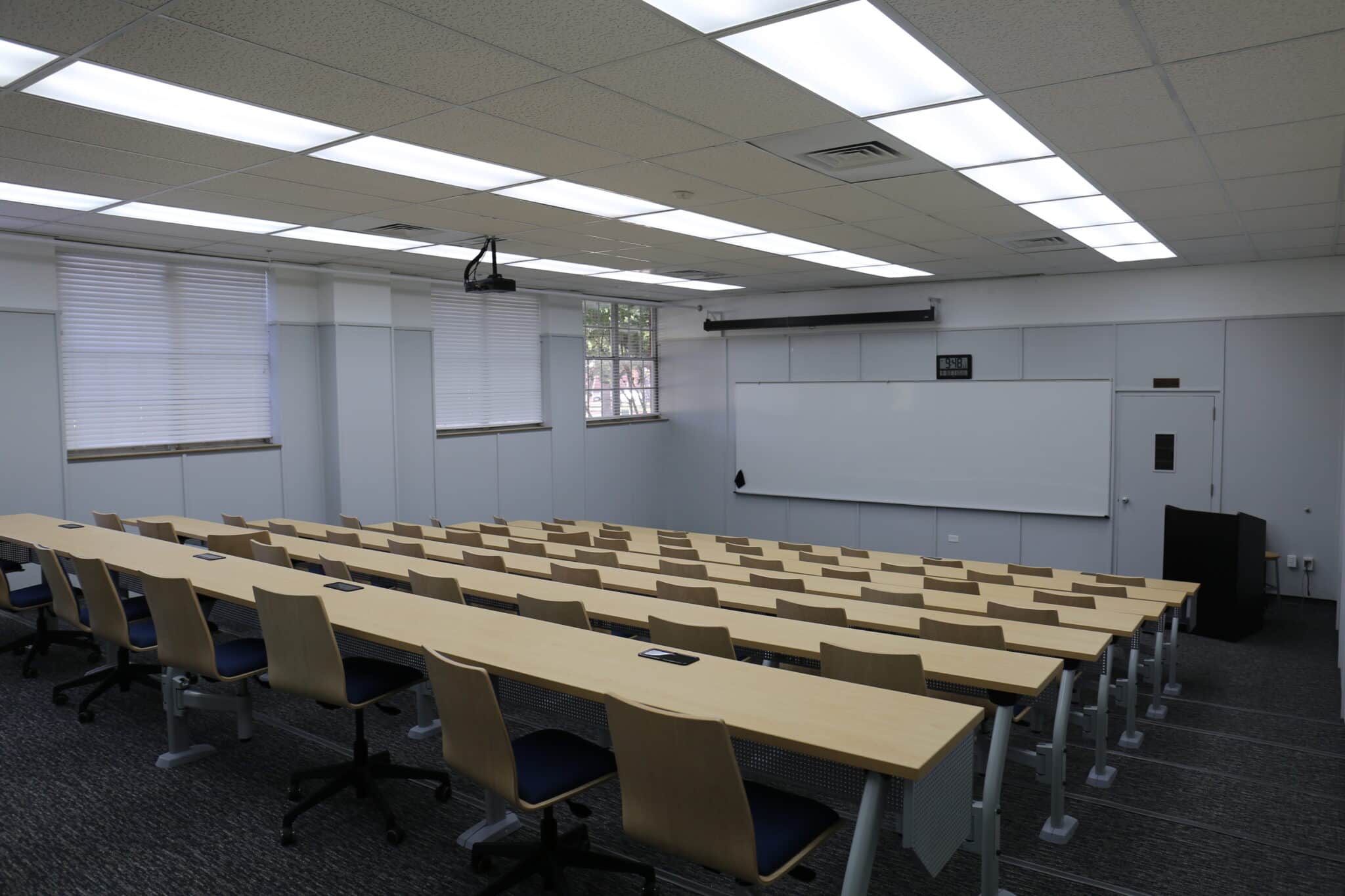 A spacious classroom at Howard Payne University with rows of tables and chairs facing a whiteboard and projector screen. | HPU
