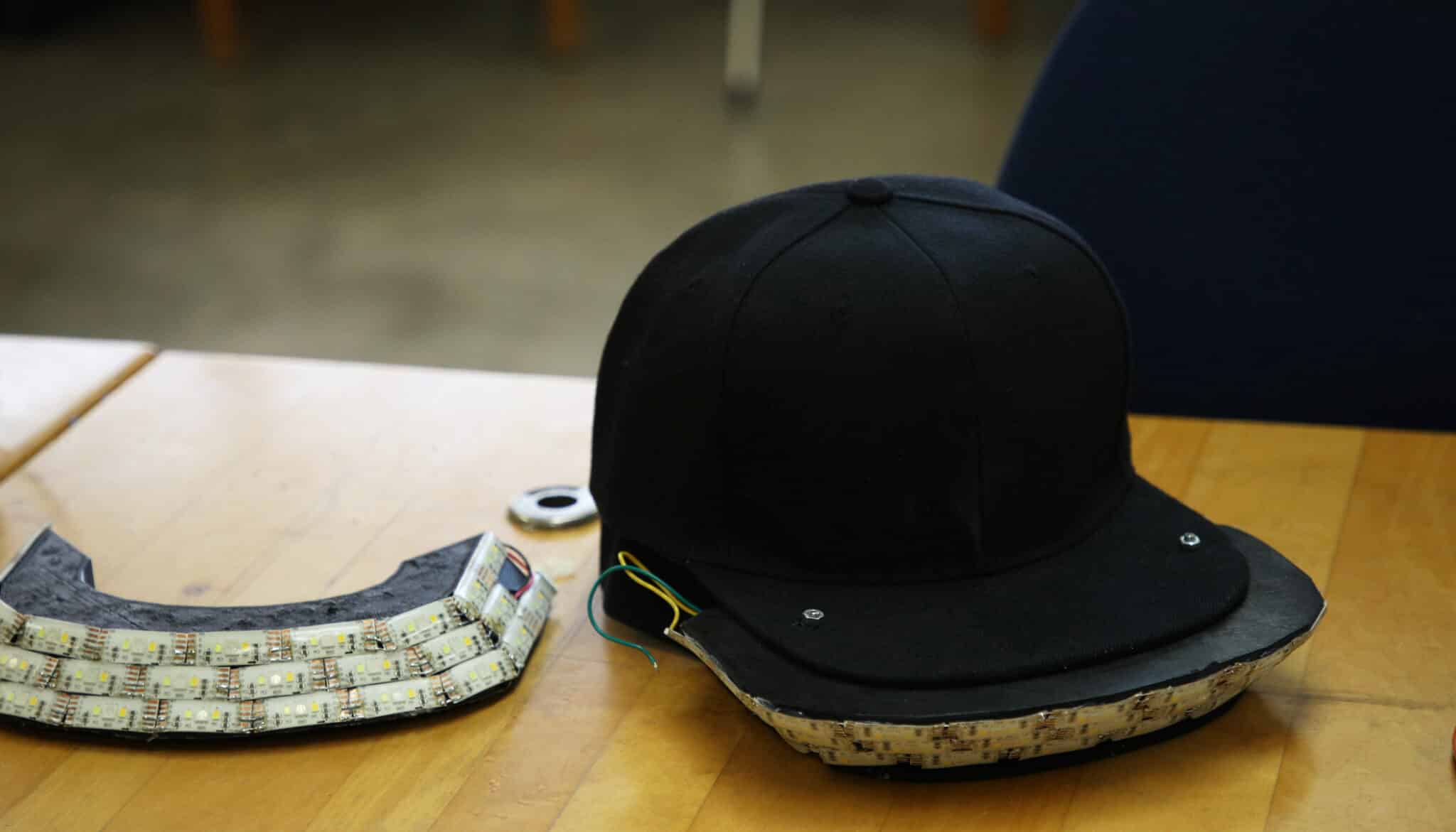 A black baseball cap with Howard Payne University logo and electronic components visible on the inner side, resting on a wooden surface in front of a chair. | HPU