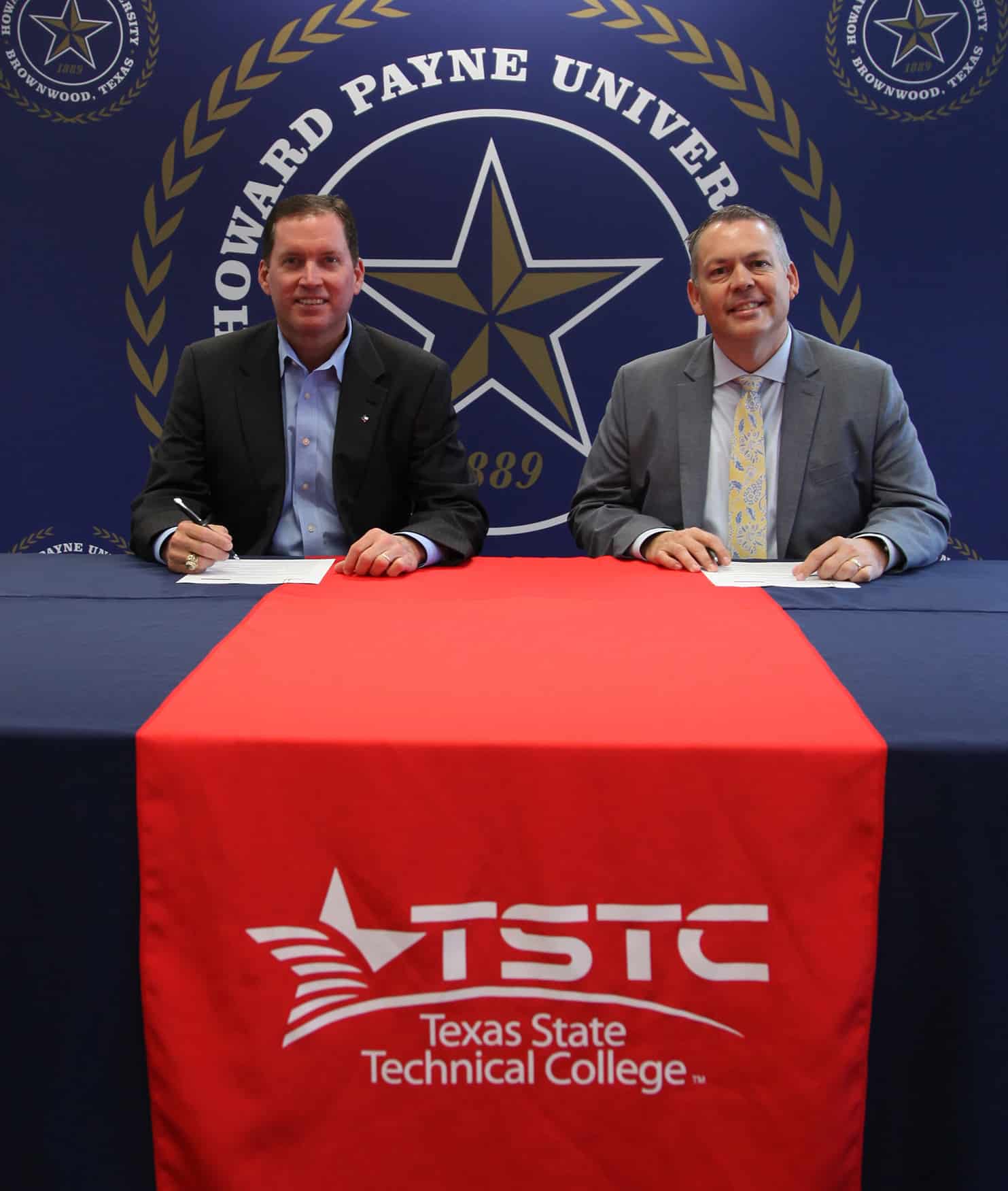 Two men in business attire sitting at a table during a formal event, possibly a signing ceremony, with banners for Howard Payne University in the background. | HPU