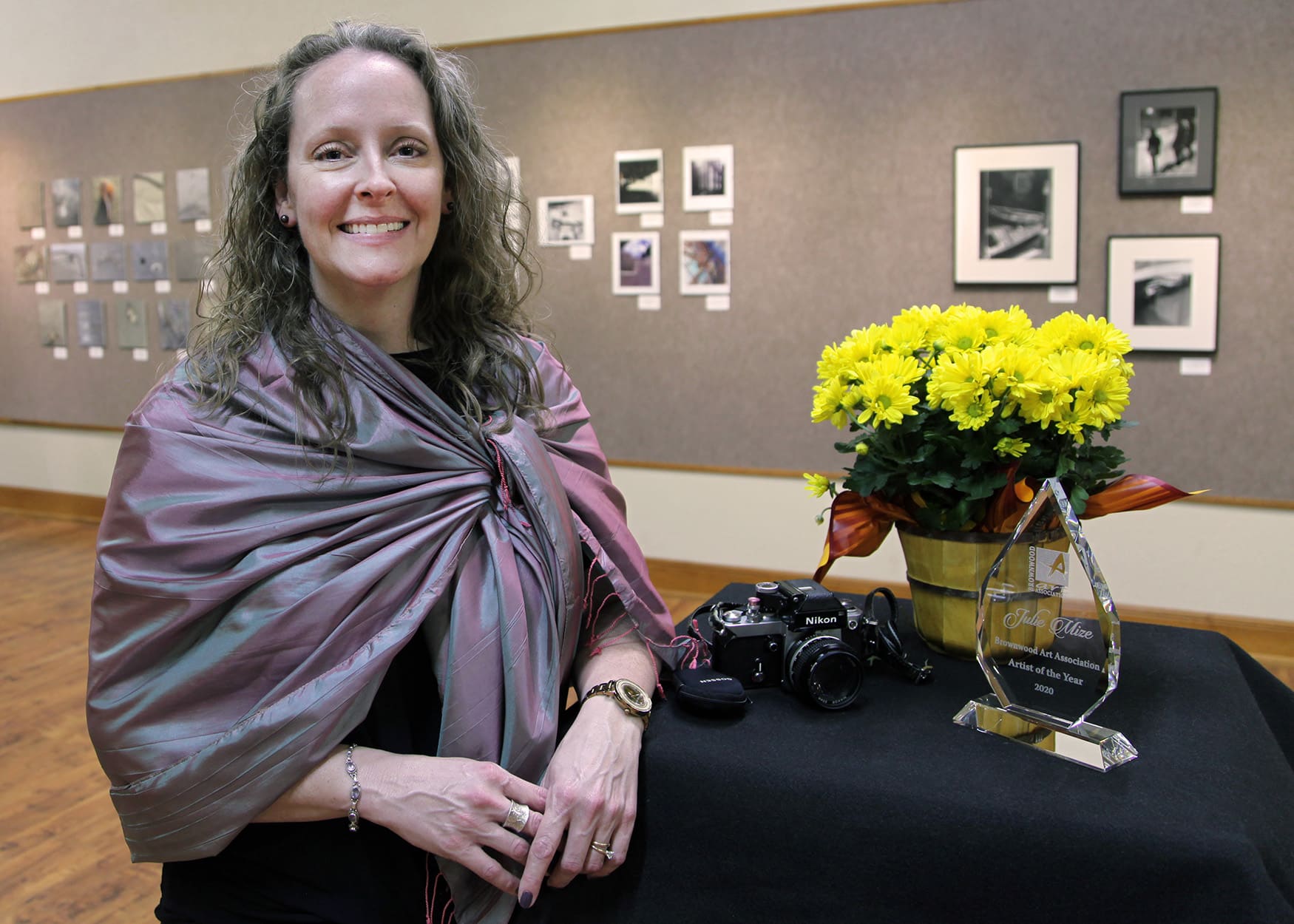 A smiling woman standing next to a camera, an award, and a bouquet of yellow flowers on a black-draped table at Howard Payne University's photography exhibition. | HPU
