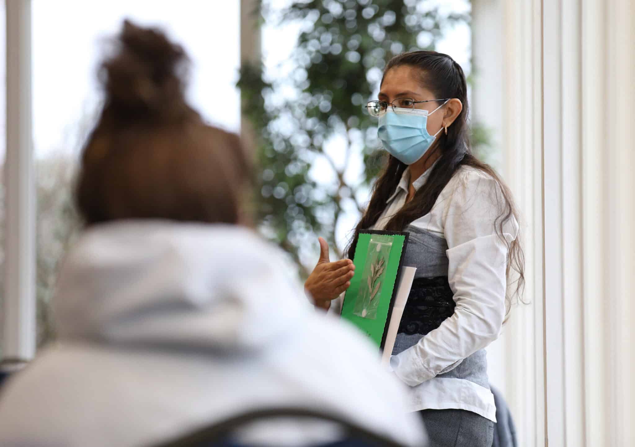 Woman wearing a face mask standing with a clipboard in a room at Howard Payne University with another person sitting in the foreground. | HPU