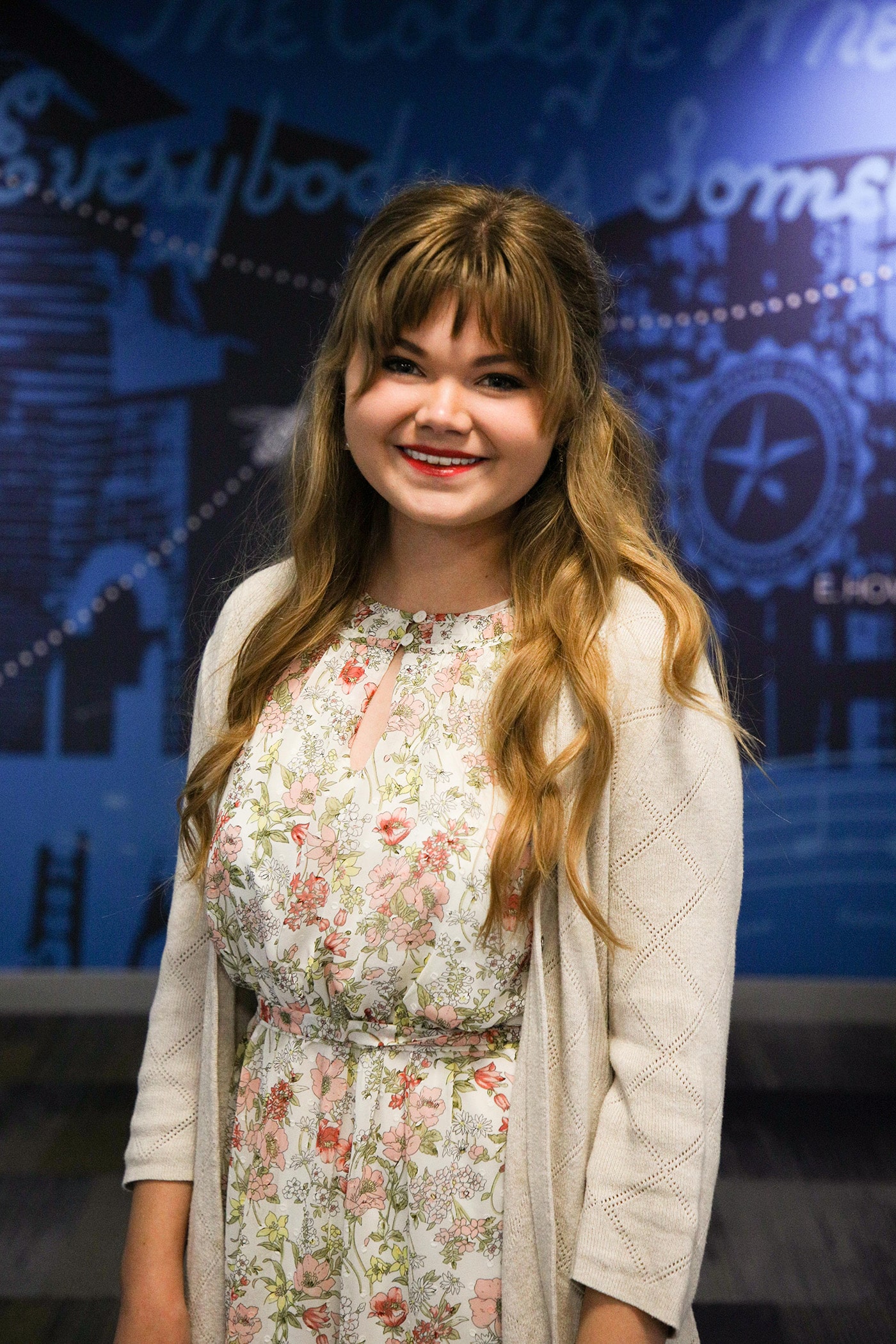 A smiling young woman in a floral dress and beige cardigan standing in front of a Howard Payne University wall with graphic designs. | HPU