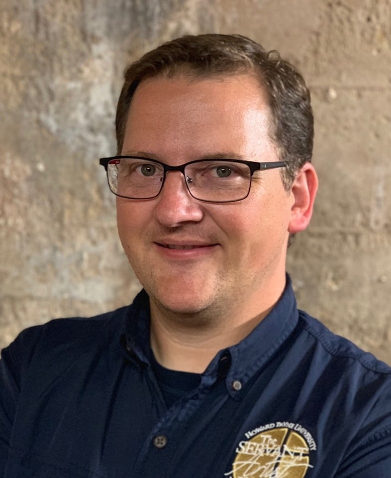 A smiling man with glasses wearing a dark blue Howard Payne University shirt against a textured background. | HPU
