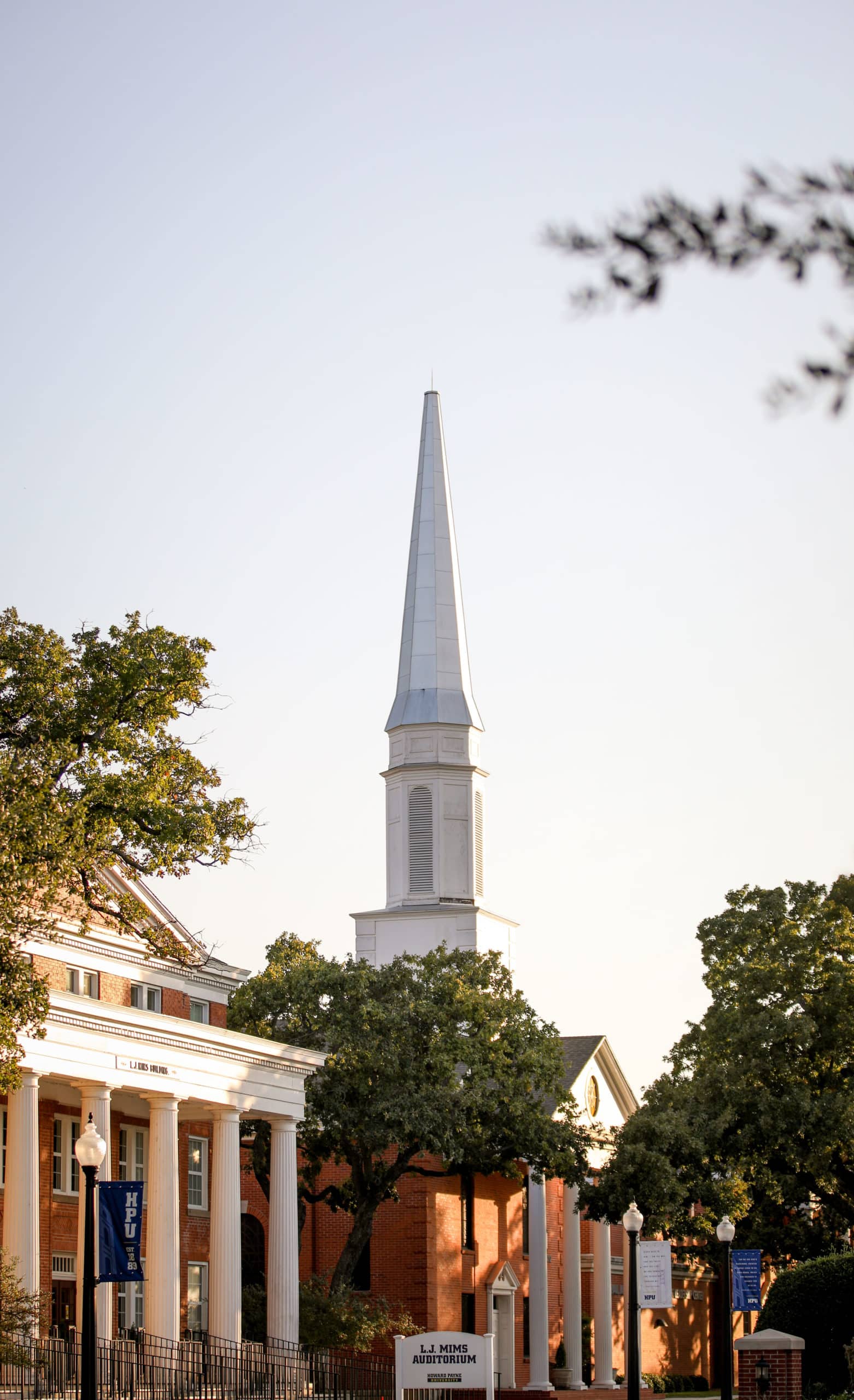A Howard Payne University church steeple rises behind a classical building with columns, bathed in warm sunlight. | HPU
