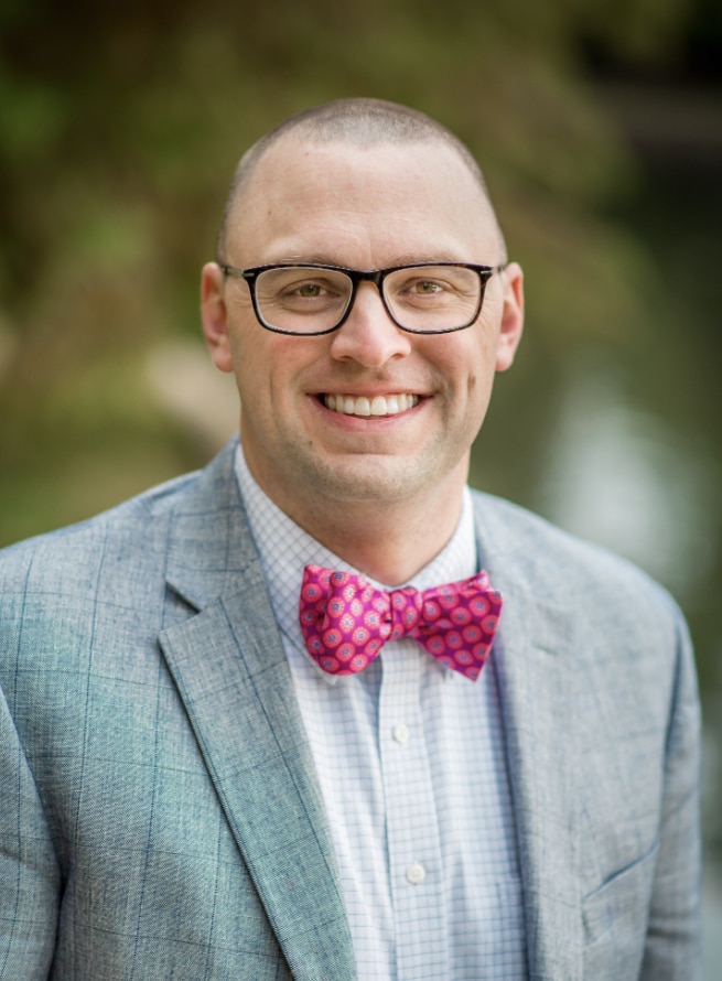 A man in glasses, wearing a patterned bow tie and a light blue blazer with Howard Payne University insignia, smiling at the camera. | HPU