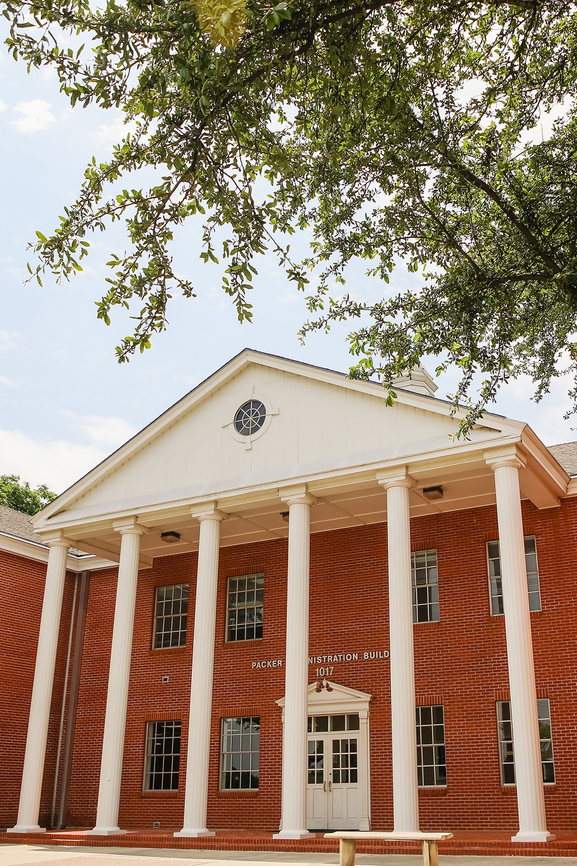 Classical red brick building with white columns and a pediment, labeled "Howard Payne University - Administration Building 1912", framed by leafy tree branches. | HPU
