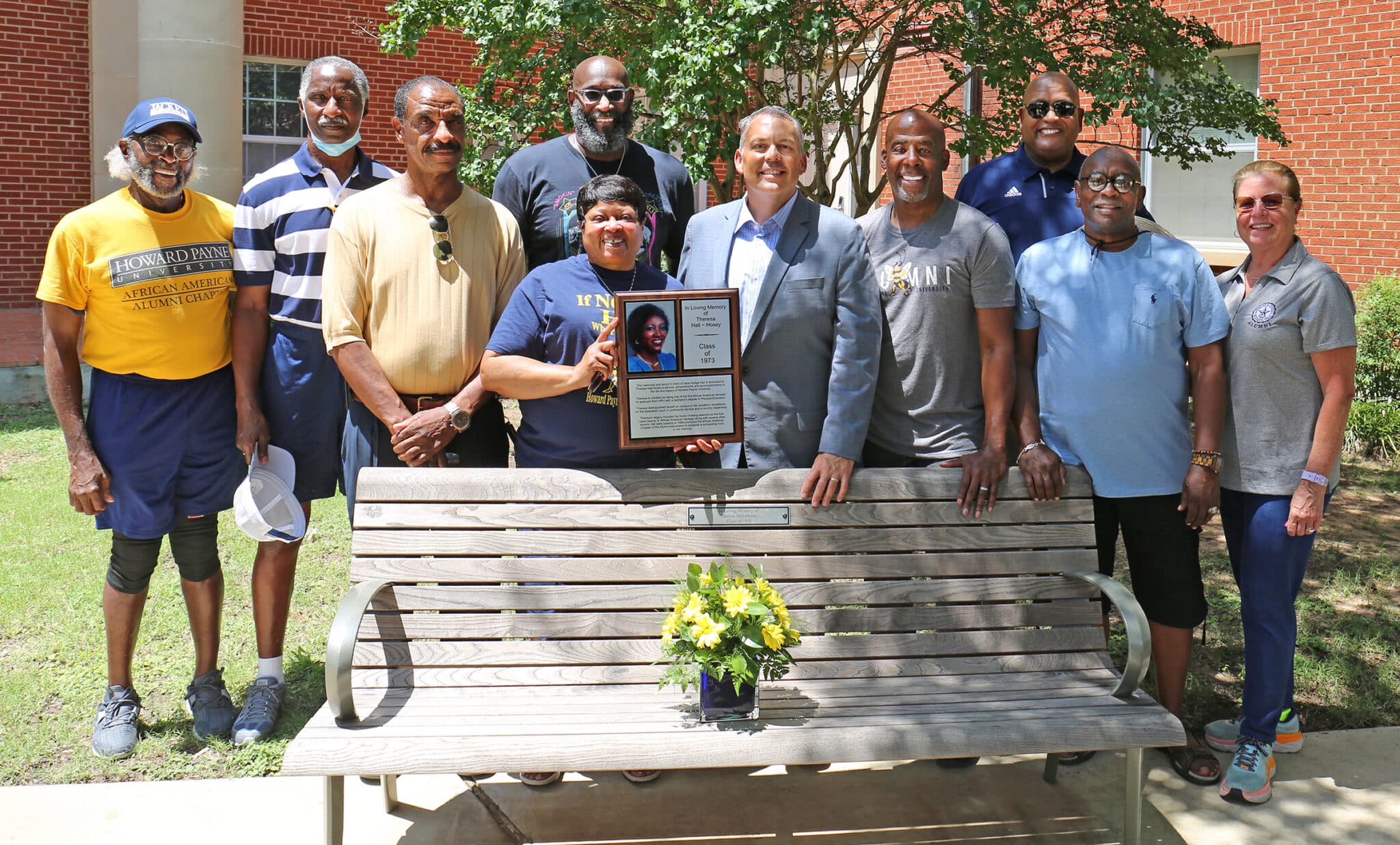 Group of people from Howard Payne University posing proudly with a commemorative plaque beside a bench. | HPU