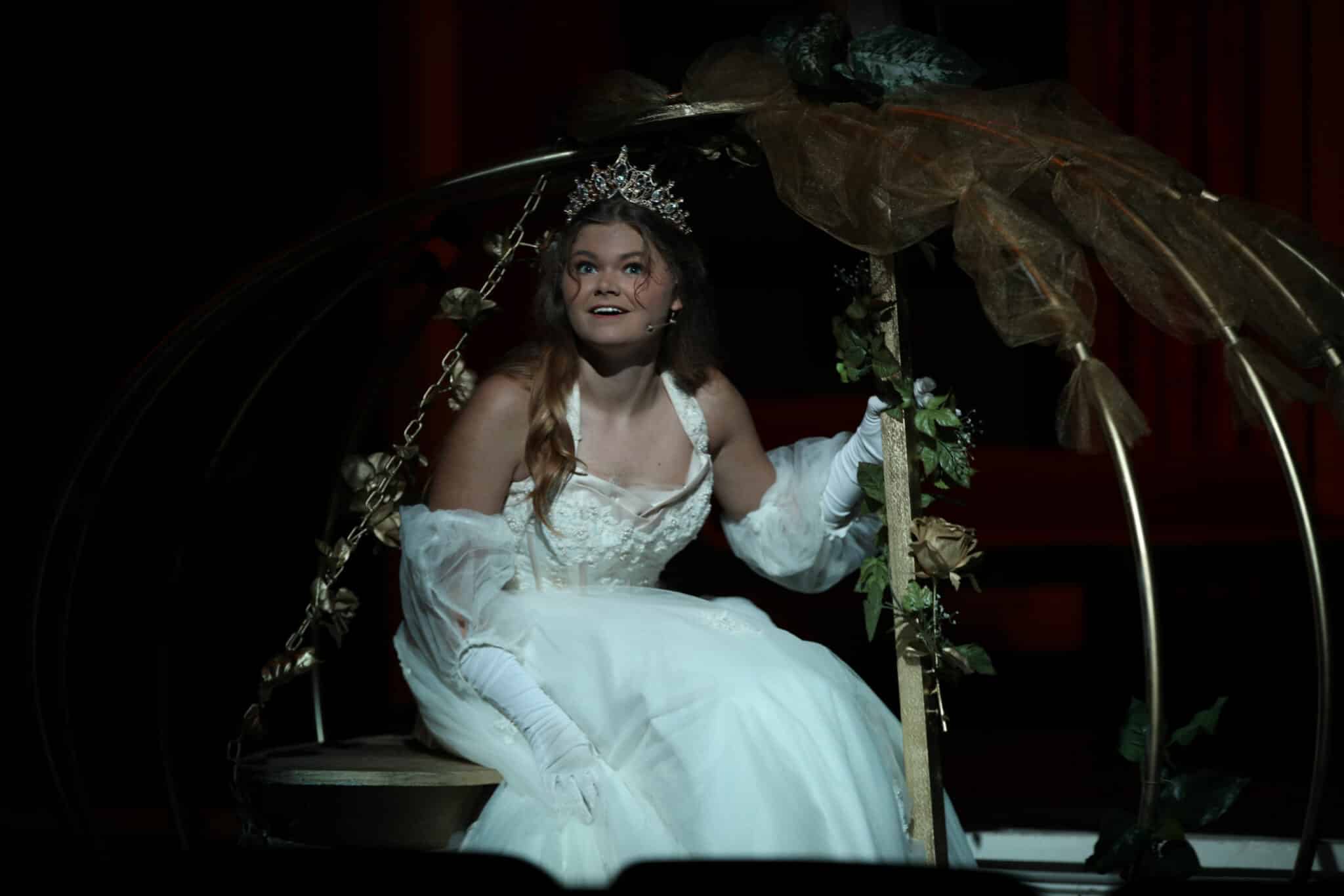 A person in a white dress and tiara sits on a swing adorned with leaves on a dark stage at Howard Payne University. | HPU
