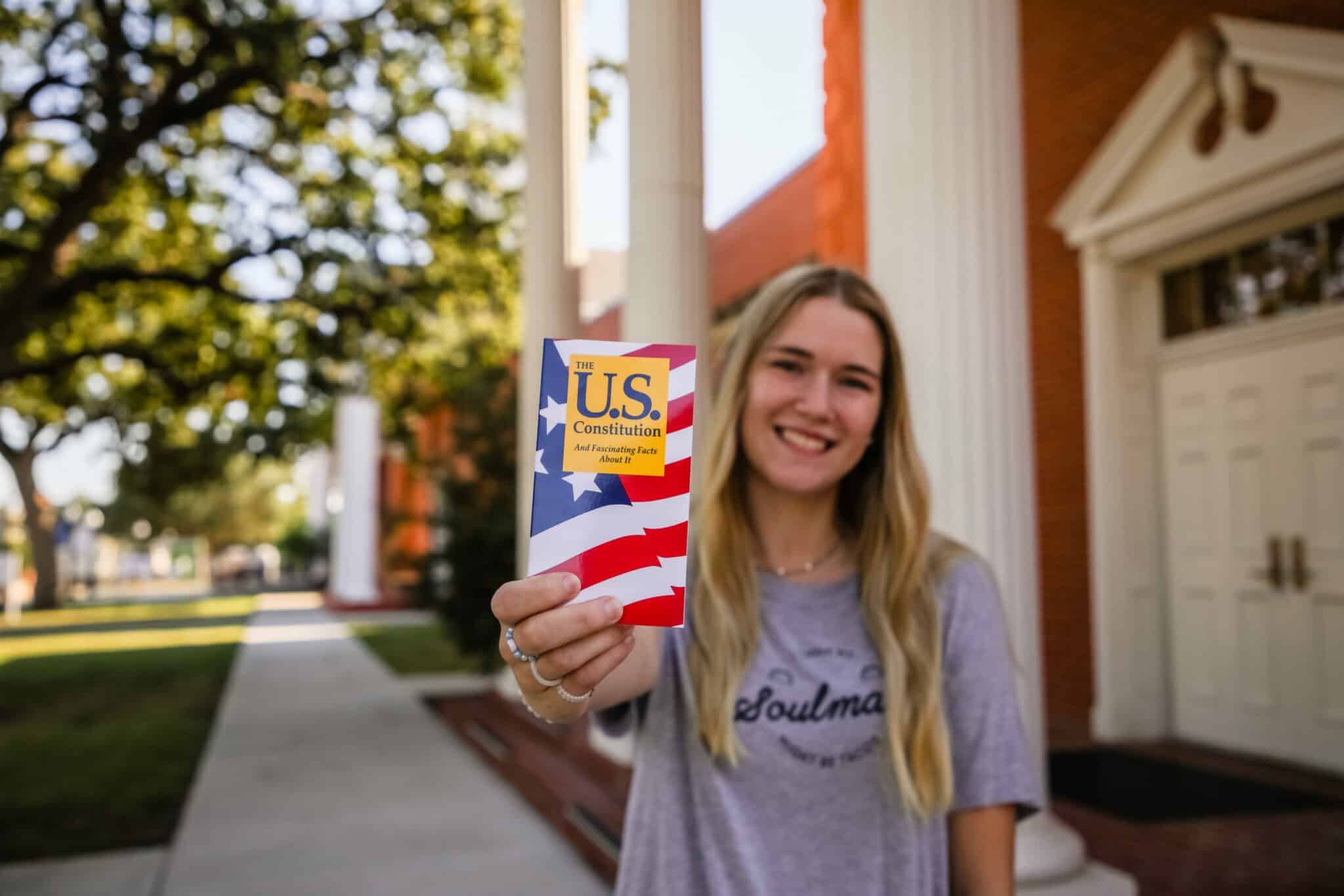 Young woman smiling and holding a U.S. Constitution booklet outdoors at Howard Payne University. | HPU