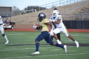 Football player from Howard Payne University in blue and gold uniform running with the ball while an opponent in white reaches to tackle him on the field. | HPU