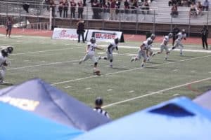 American football game in progress with a Howard Payne University player kicking off the ball as teams advance on the field. | HPU
