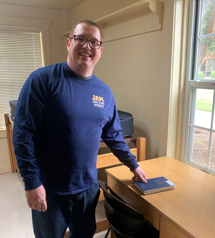 A smiling man standing next to a desk with a Howard Payne University book on it. | HPU