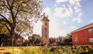 A sunlit Howard Payne University campus with a clock tower, surrounded by trees and buildings under a clear sky. | HPU