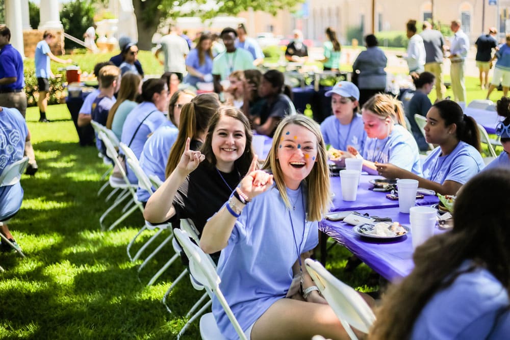 Group of Howard Payne University people wearing blue shirts enjoying an outdoor gathering with food and drinks on a sunny day. | HPU