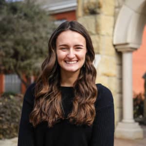 A smiling woman with long wavy hair, wearing a black sweater, standing in front of Howard Payne University's building with stone arches. | HPU