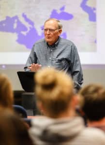 A lecturer at Howard Payne University speaking to a classroom of students with a world map in the background. | HPU