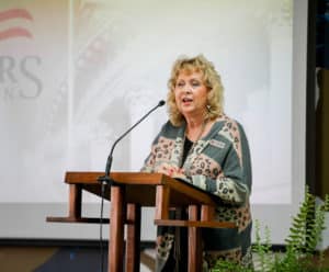 A woman speaking at a podium with a microphone in a room with an American flag and the Howard Payne University emblem in the background. | HPU