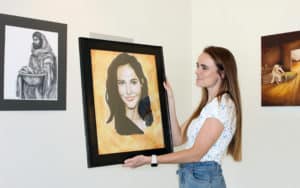 A woman hangs a framed portrait on the wall at a Howard Payne University art exhibition. | HPU