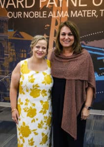 Two women posing for a photo at a Howard Payne University event, one in a floral dress and the other in a black outfit with a brown shawl. | HPU
