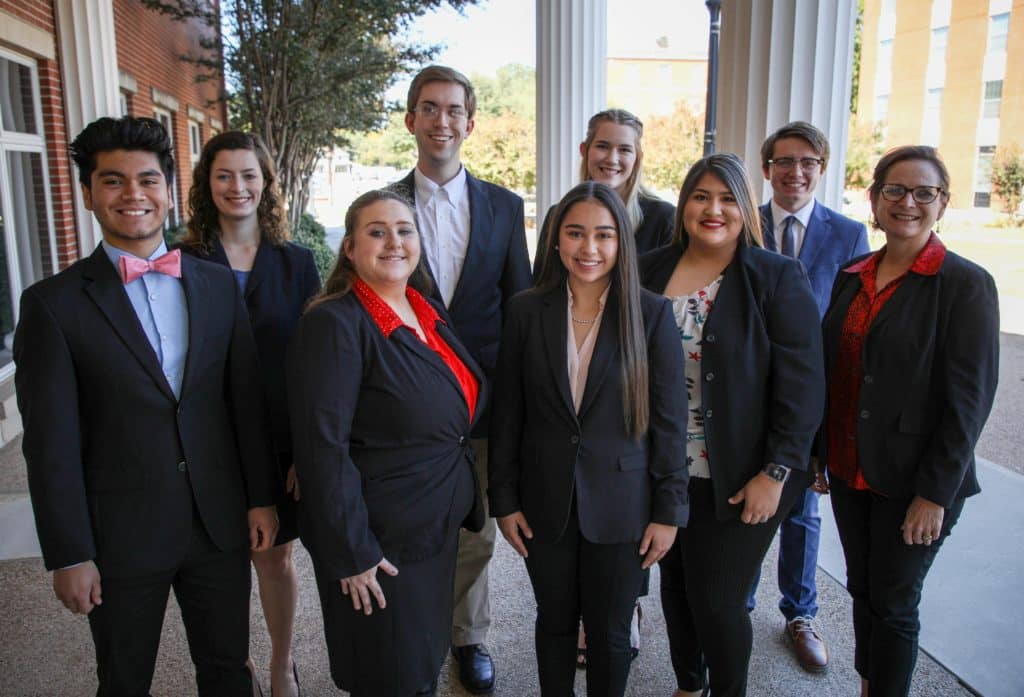 Group of eight well-dressed individuals smiling for a photo outside a building with columns at Howard Payne University. | HPU