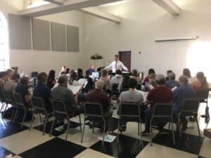 Conductor leading a Howard Payne University choir rehearsal in a room with seated singers. | HPU