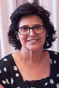 A woman with short dark hair and glasses, wearing a black dress with daisy patterns and Howard Payne University insignia, smiling at the camera. | HPU
