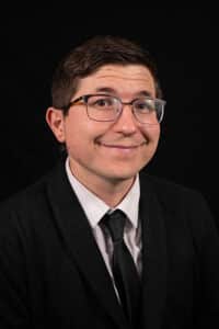 A man in a Howard Payne University suit and tie smiling at the camera against a black background. | HPU