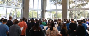 A group of people gathered in a room with large windows at Howard Payne University, attentively facing a speaker at the front. | HPU
