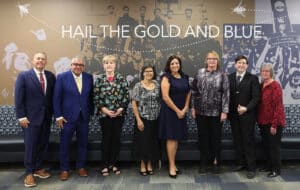 A group of seven adults standing in front of a wall with the inscription "hail the gold and blue," possibly at a formal event or gathering at Howard Payne University. | HPU