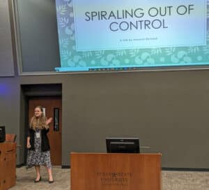 A woman gesturing with her hand stands beside a podium in a lecture hall, with a presentation titled "spiraling out of control" on the screen. | HPU