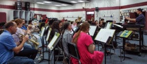 A band rehearsal in a music room with a conductor leading diverse musicians playing various instruments, including brass, woodwinds, and percussion. | HPU